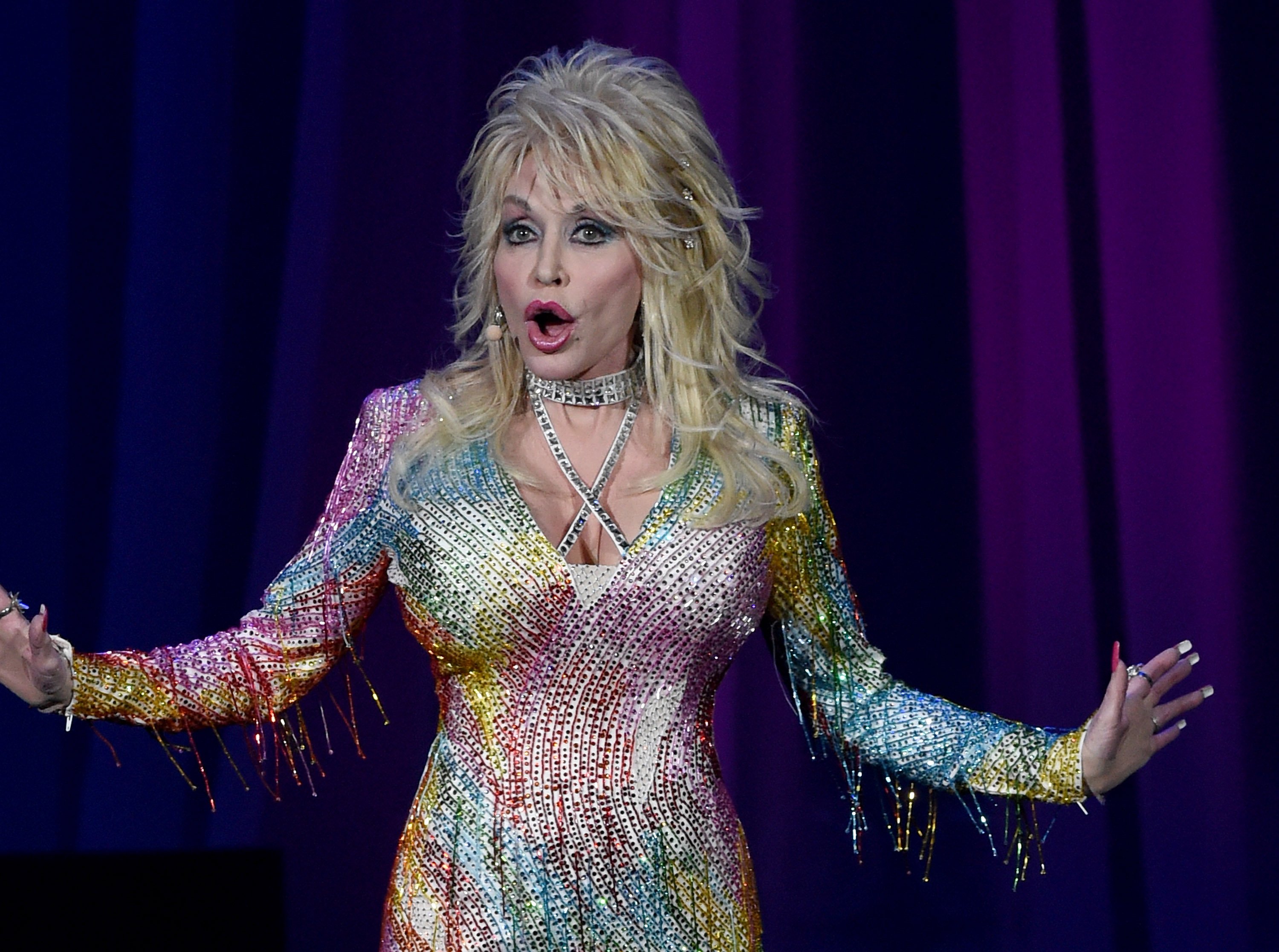 Dolly Parton singing on stage in a rainbow dress and blonde wig.