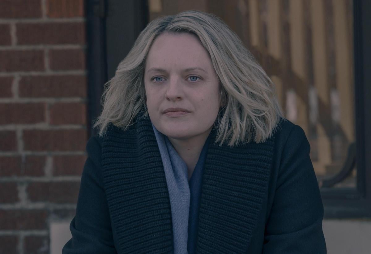 Elisabeth Moss sits on a stoop wearing a light blue scarf and black coat as June in 'The Handmaid's Tale' Season 4.