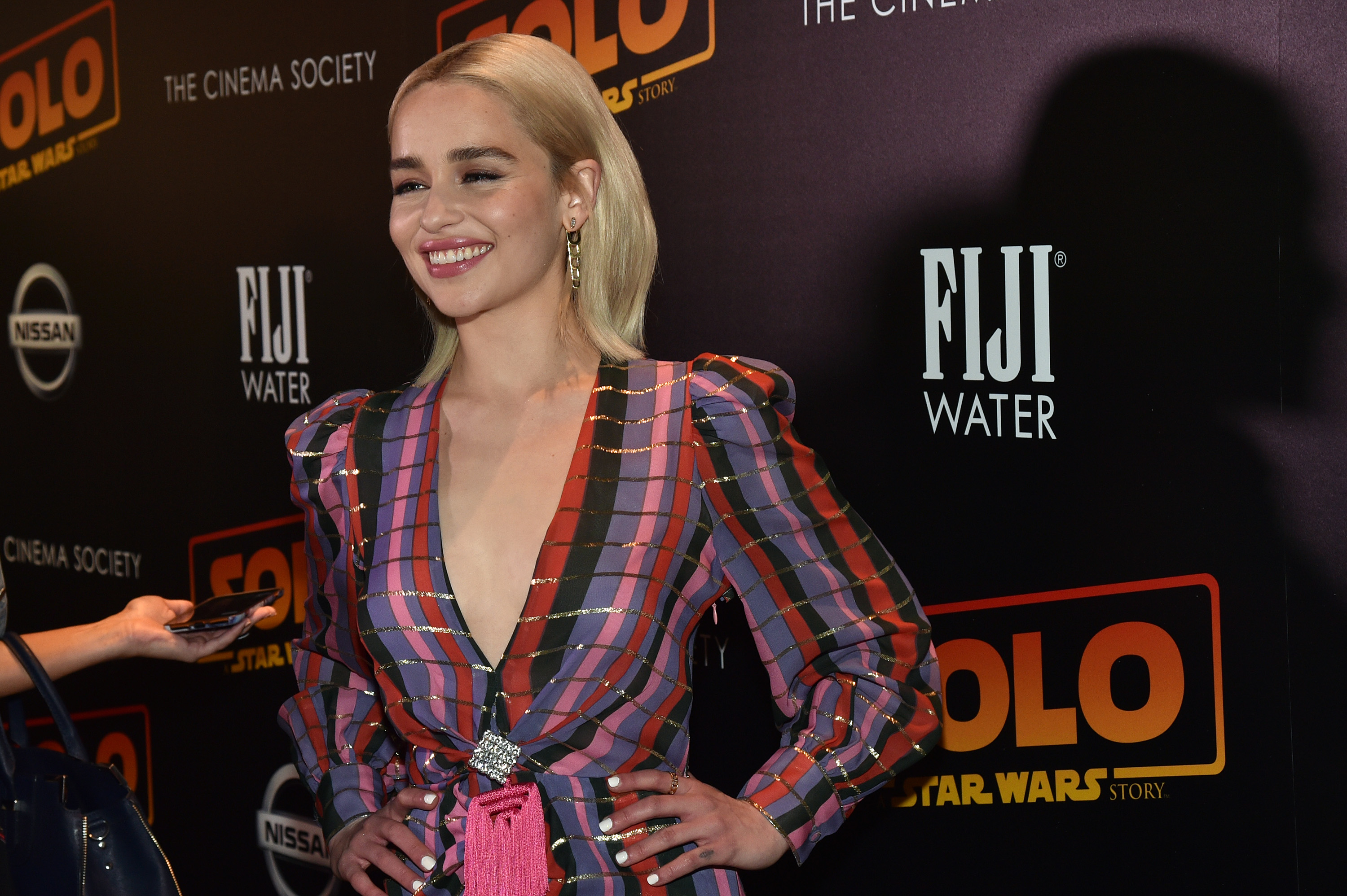 'Solo' star Emilia Clarke wearing a pink, purple, and red dress and standing in front of a 'Solo' and FIJI wall