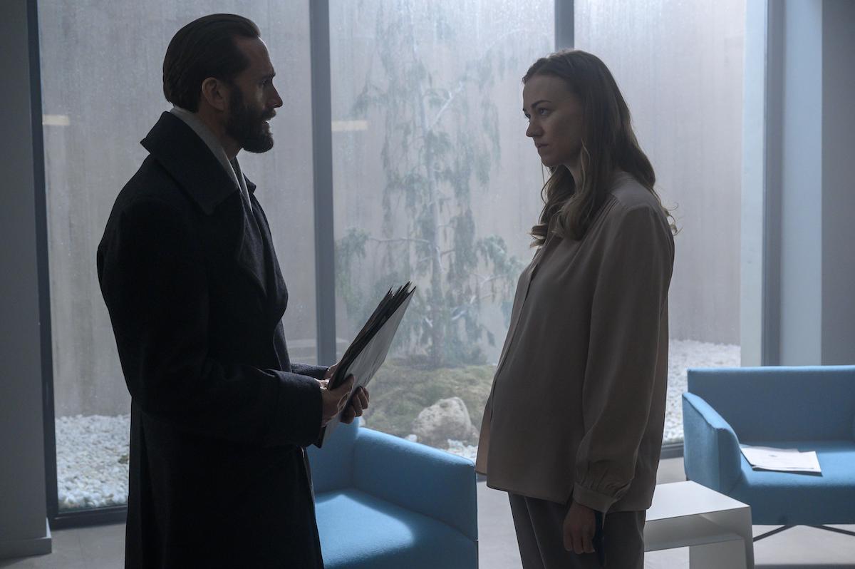 Joseph Fiennes as Fred Waterford in a long, black, wool coat holding a folder of paperwork. He stands across from Yvonne Strahovski as Serena Joy Waterford, who is pregnant and wears a light pink blouse and light grey pants in 'The Handmaid's Tale' Season 4. They stand in an elite prison cell with floor-to-ceiling windows and blue chairs. Natural light is coming through the windows and a small tree is seen through it.