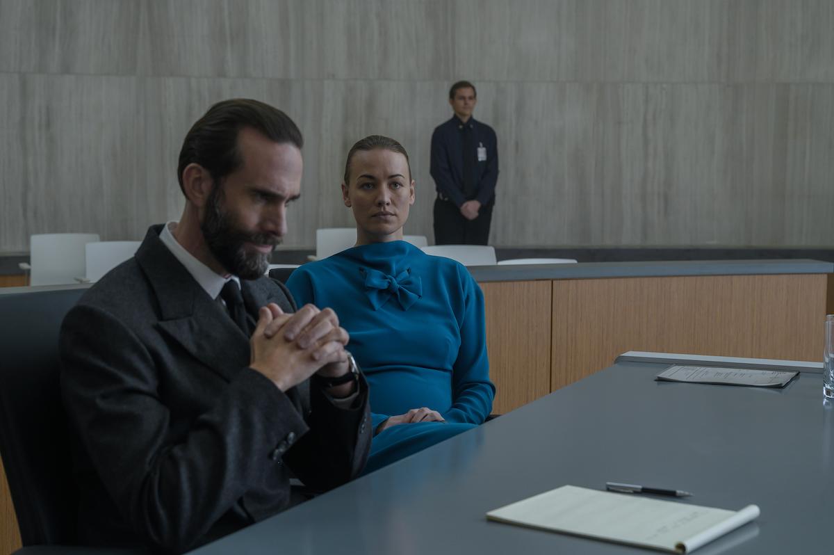Joseph Fiennes as Fred Waterford and Yvonne Strahovski as Serena Joy Waterford in 'The Handmaid's Tale' Season 4. He wears a dark grey suit and crosses his hands in concern while sitting at a table in a court room. She wears a teal dress and looks straight ahead while sitting next to him.