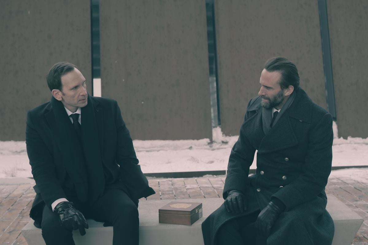 Stephen Kunken as Warren Putnam and Joseph Fiennes as Fred Waterford in matching suits and coats sitting outside on a concrete bench in winter in 'The Handmaid's Tale' Season 4