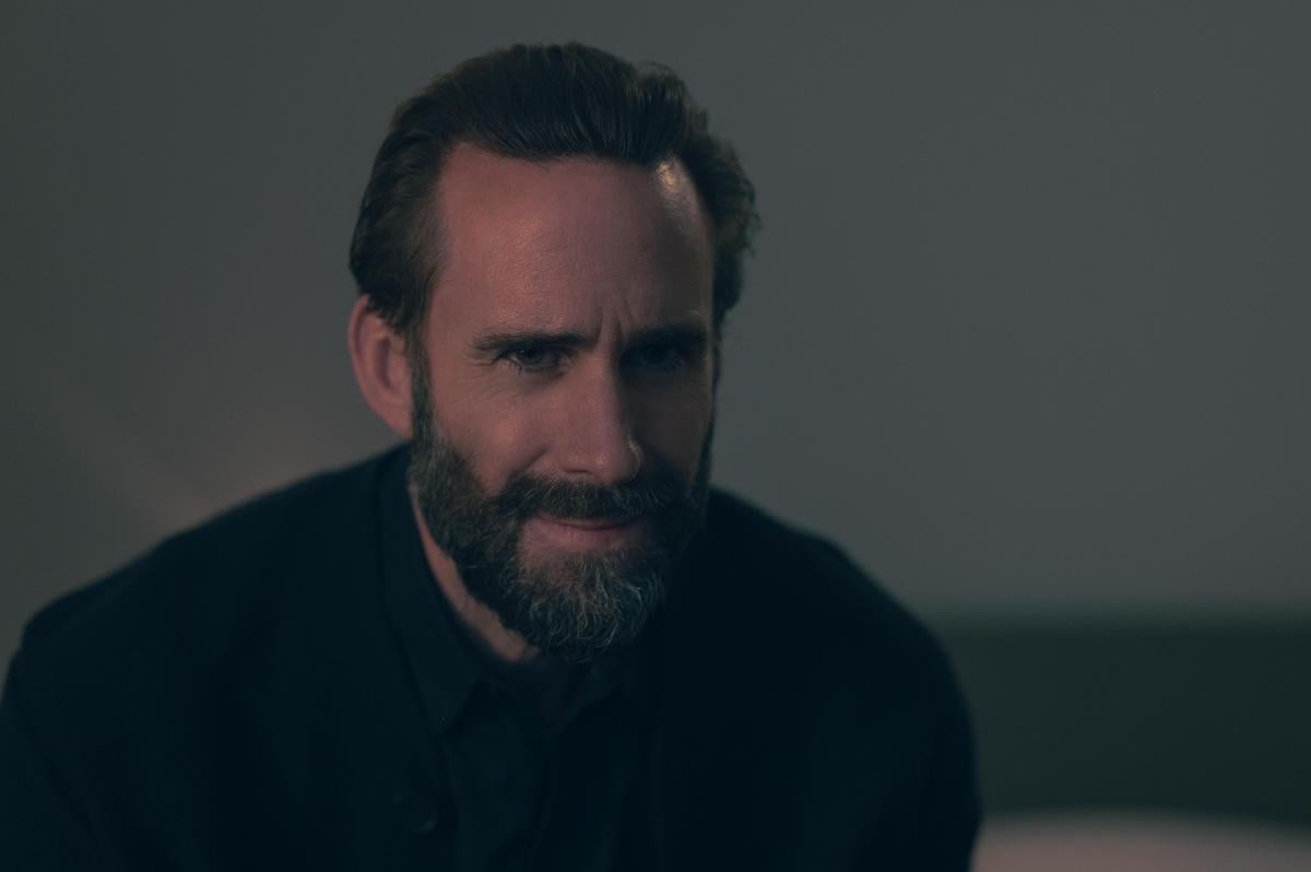 Joseph Fiennes as Fred Waterford wearing a black shirt, black sweater, and black tie in front of a beige wall in 'The Handmaid's Tale' Season 4