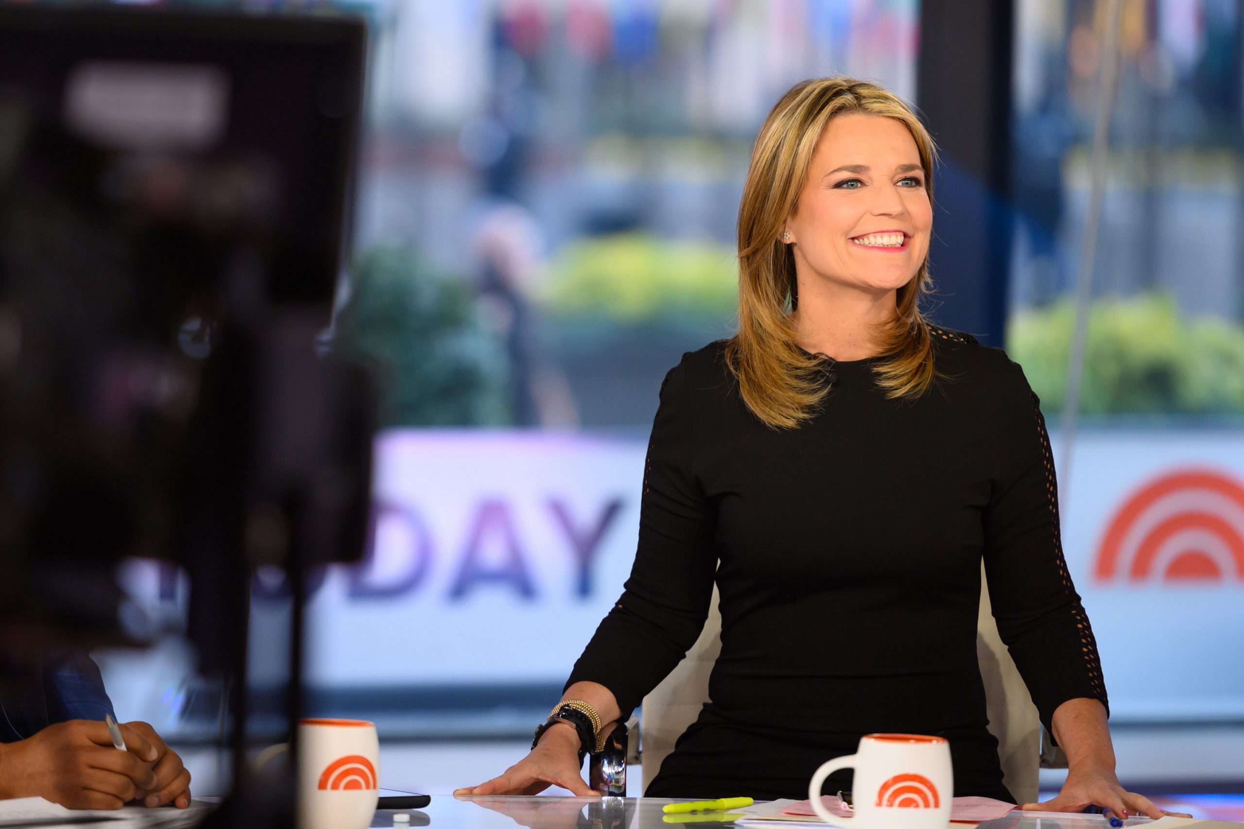 Morning show co-host Savannah Guthrie smiles as she sits at her anchor chair on the set of 'Today' in a long-sleeved black dress.