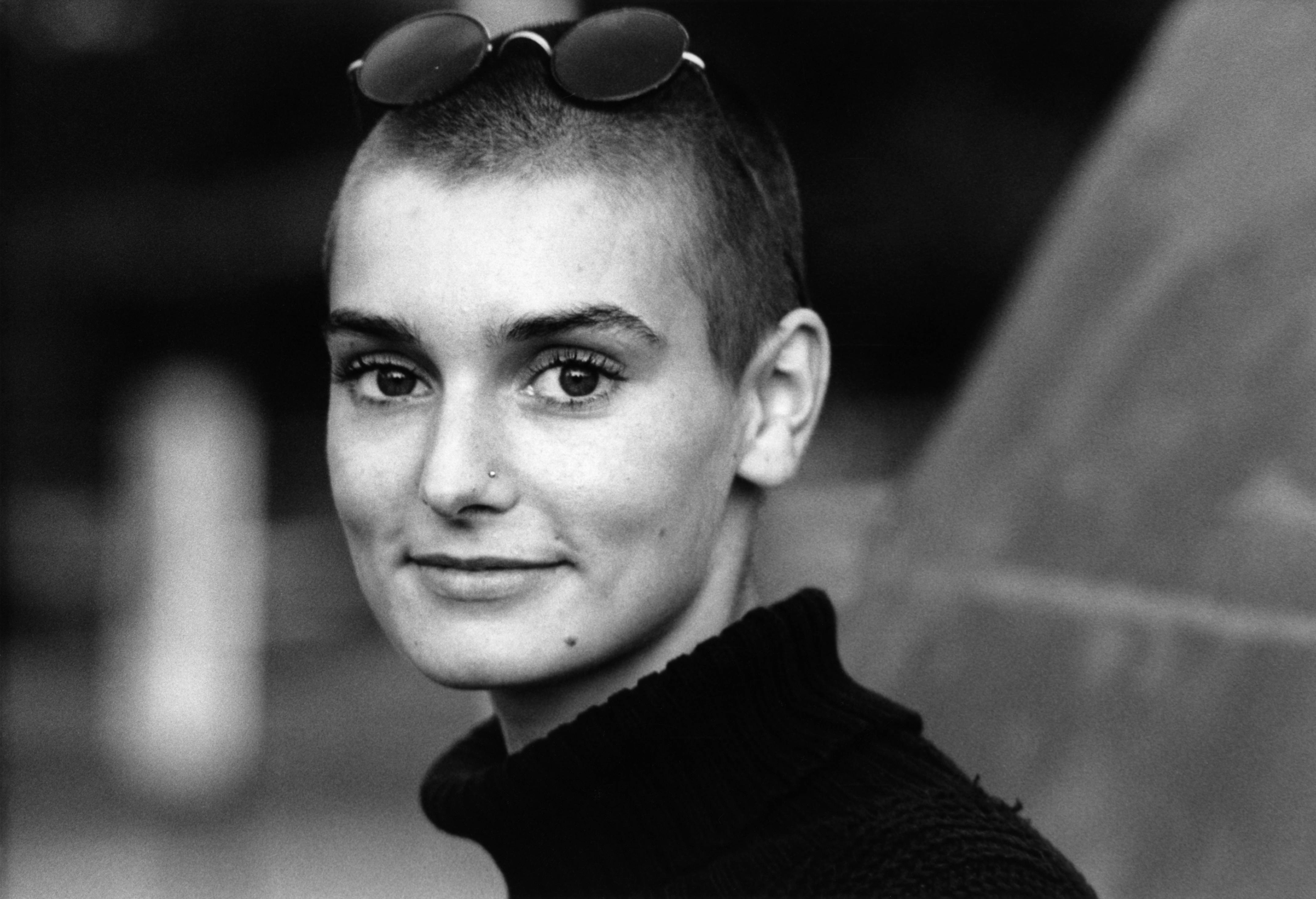 With sunglasses perched on her head, Irish singer Sinéad O'Connor wears a black turtleneck and grins in this 1990 photo.