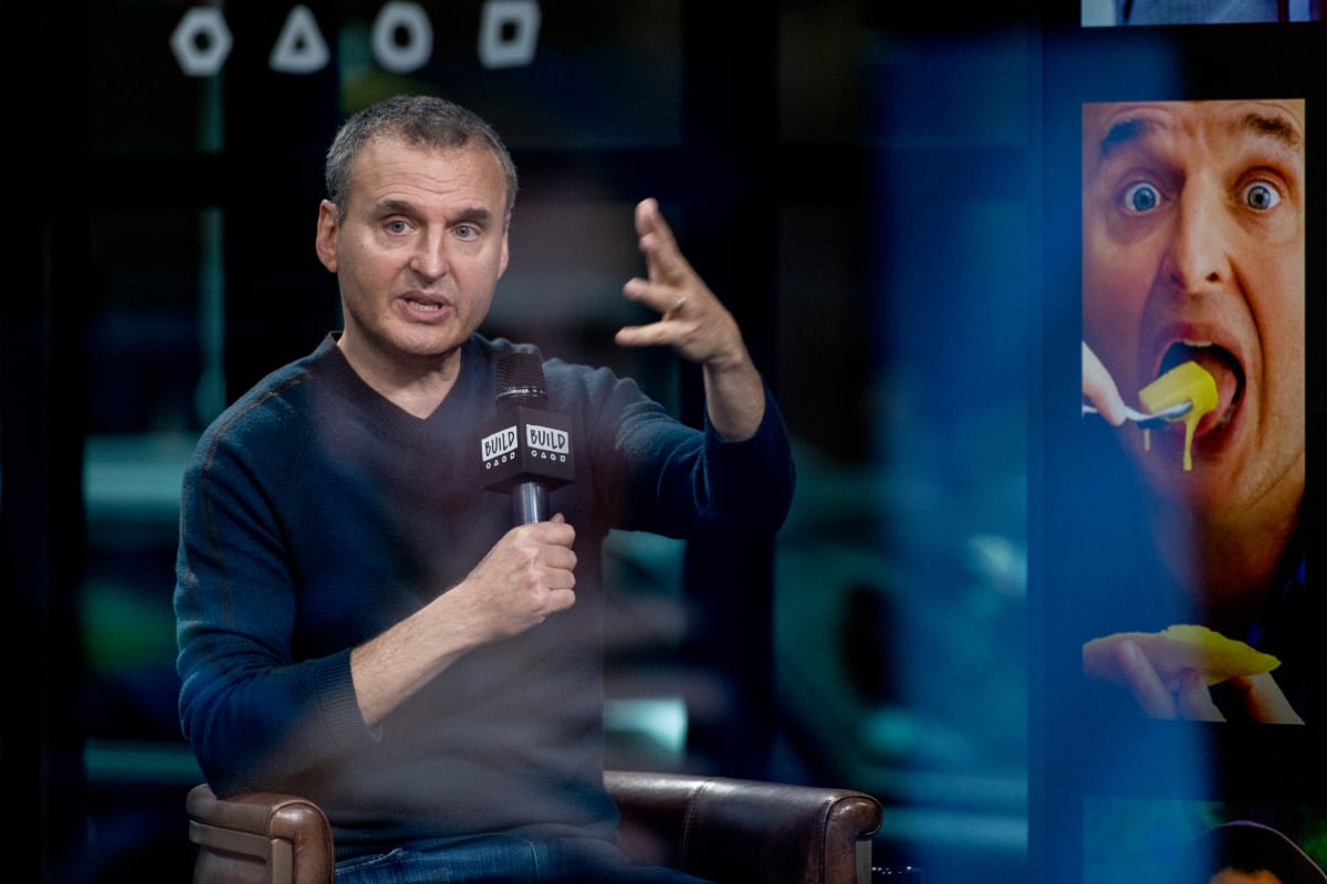 'Somebody Feed Phil' star Phil Rosenthal is seated and holds a microphone as he discusses his Netflix travel and food series alongside a promotional poster showing the host with a spoonful of food in front of his face.