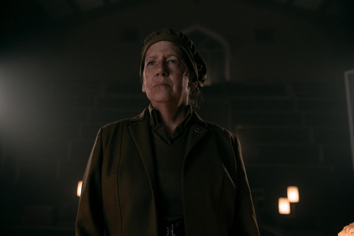 Ann Dowd as Aunt Lydia in 'The Handmaid's Tale' Season 4. She stands on a podium with a bruised face wearing a brown hat and brown dress and coat.