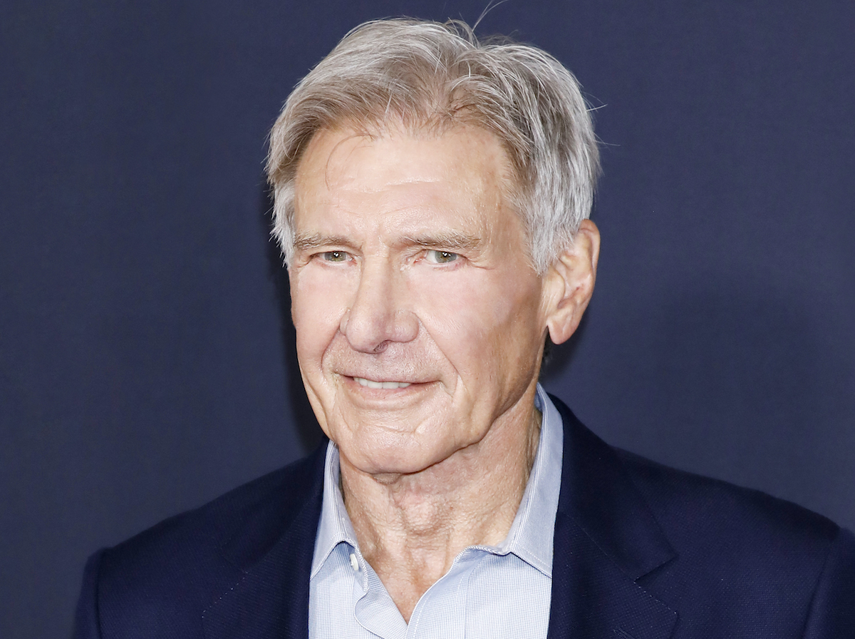 Harrison Ford smiles in a navy blue suit and light blue button-down shirt in front of a blue-grey background.