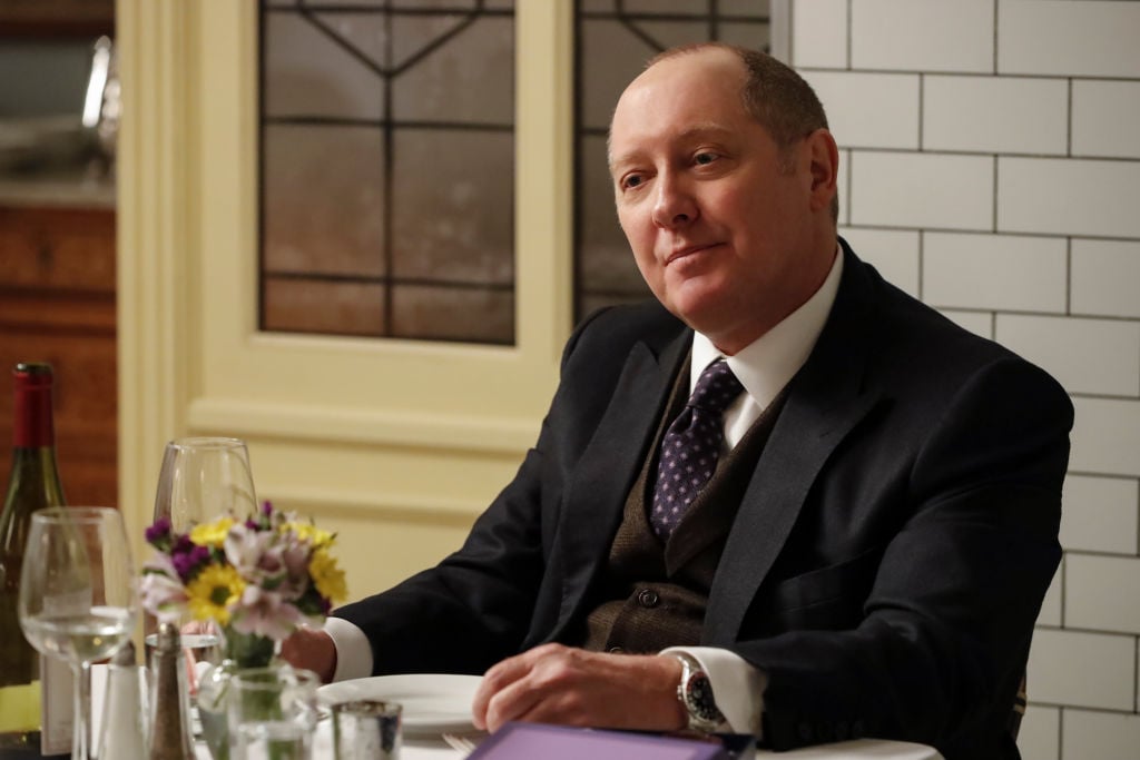 James Spader as Raymond 'Red' Reddington looks across the table he's sitting at with a smirk.