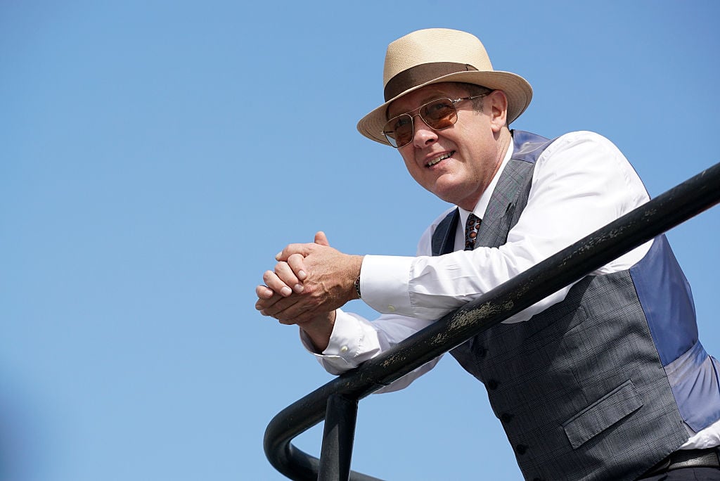 James Spader as Red Reddington stands over a railing. He's wearing sunglasses and a sun hat with a smile on his face.