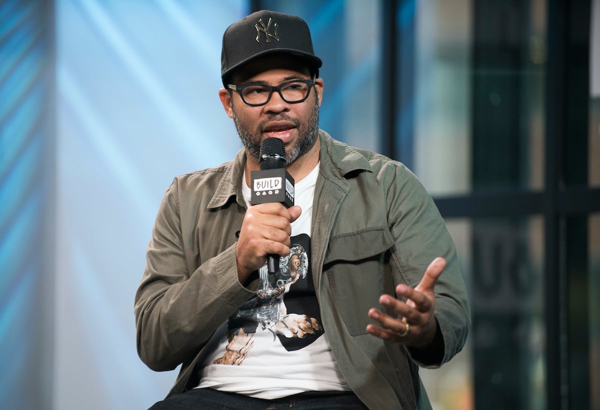 Jordan Peele wears a hat and jacket as he speaks into a microphone