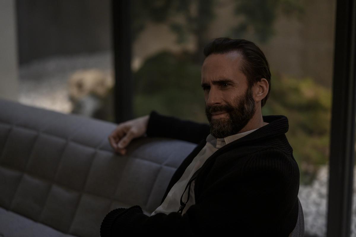 Joseph Fiennes as Fred Waterford in 'The Handmaid's Tale' Season 4. He sits on a grey couch in a high-end prison cell with floor-to-ceiling windows. He wears a white button-down shirt and black sweater, his arm is on the back of the couch.