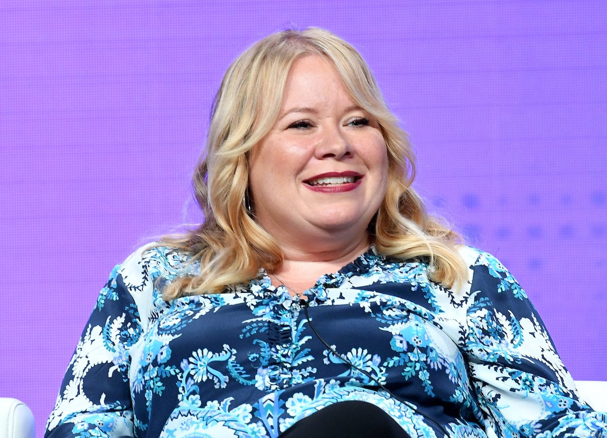 'The Vampire Diaries' creator Julie Plec smiles in a blue floral shirt while sitting in a white chair in front of a purple background.