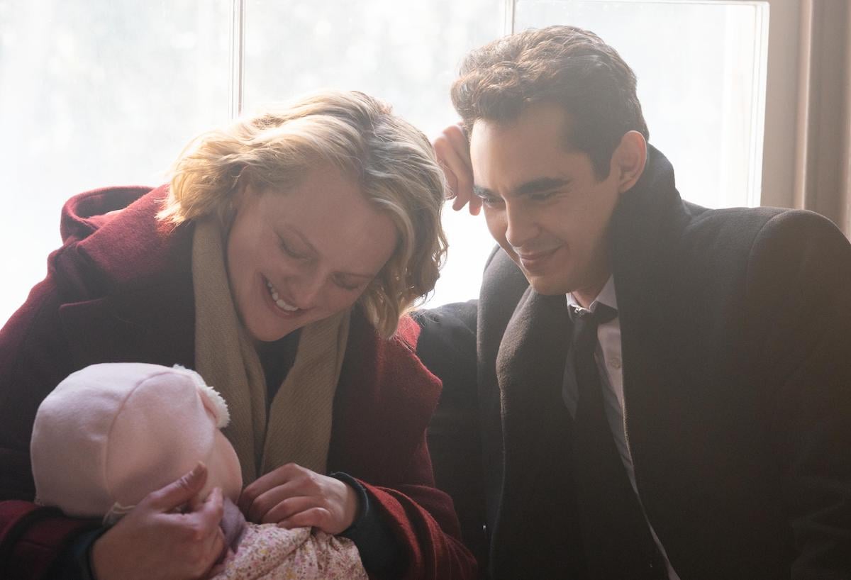 Elisabeth Moss wears a red wool coat and beige scarf as June and Max Minghella wears a black wool coat, black suit, white shirt, and black tie as Nick in 'The Handmaid's Tale' Season 4. They smile down at baby Nichole, wearing all pink. And they're sitting in front of a sun-filled window.