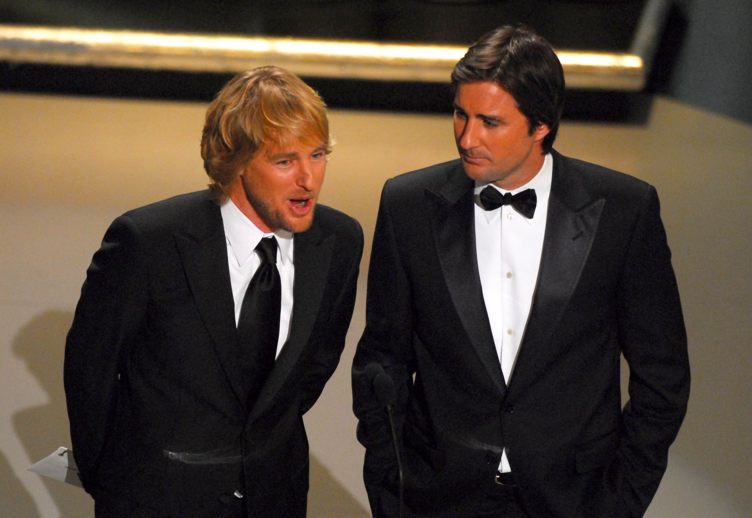 L-R: Owen Wilson and Luke Wilson in suits with Owen speaking into a microphone