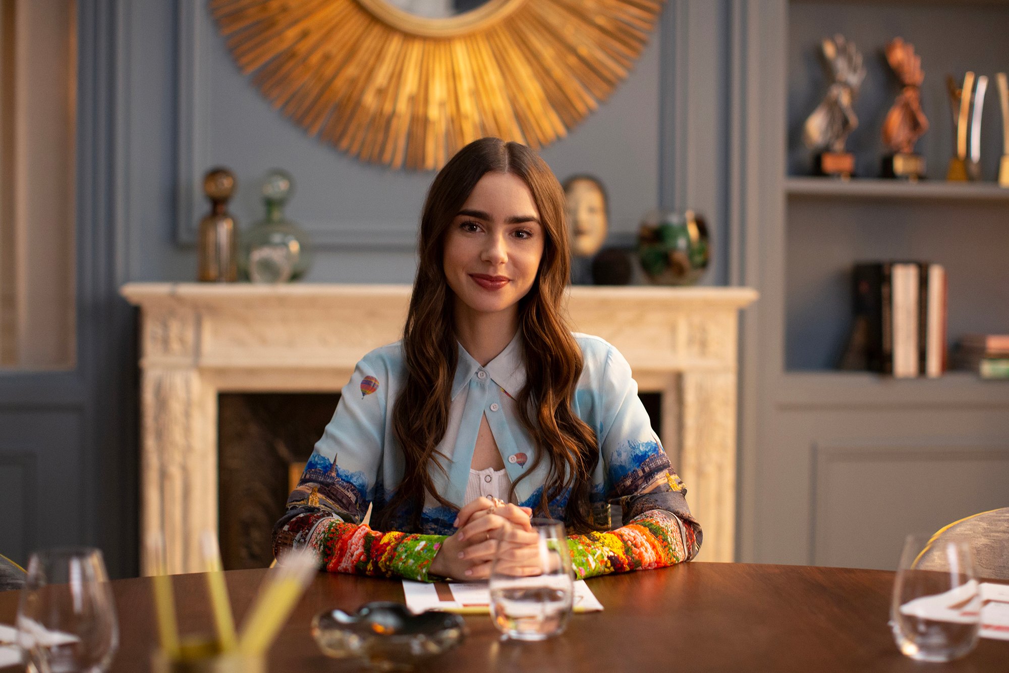 Lily Collins sits at a desk with her hands folded on 'Emily in Paris'