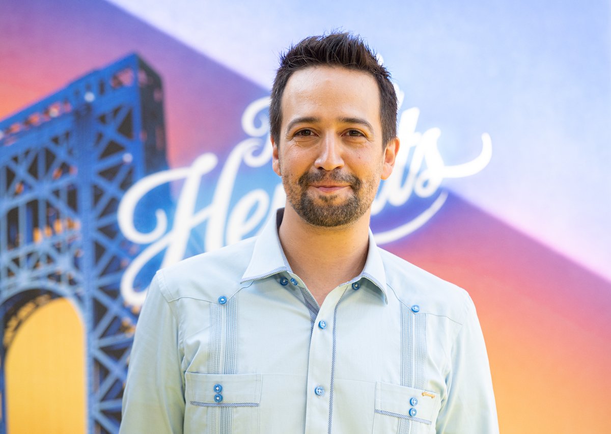 Lin-Manuel Miranda smiles in blue clothes in front of the 'In The Heights' poster