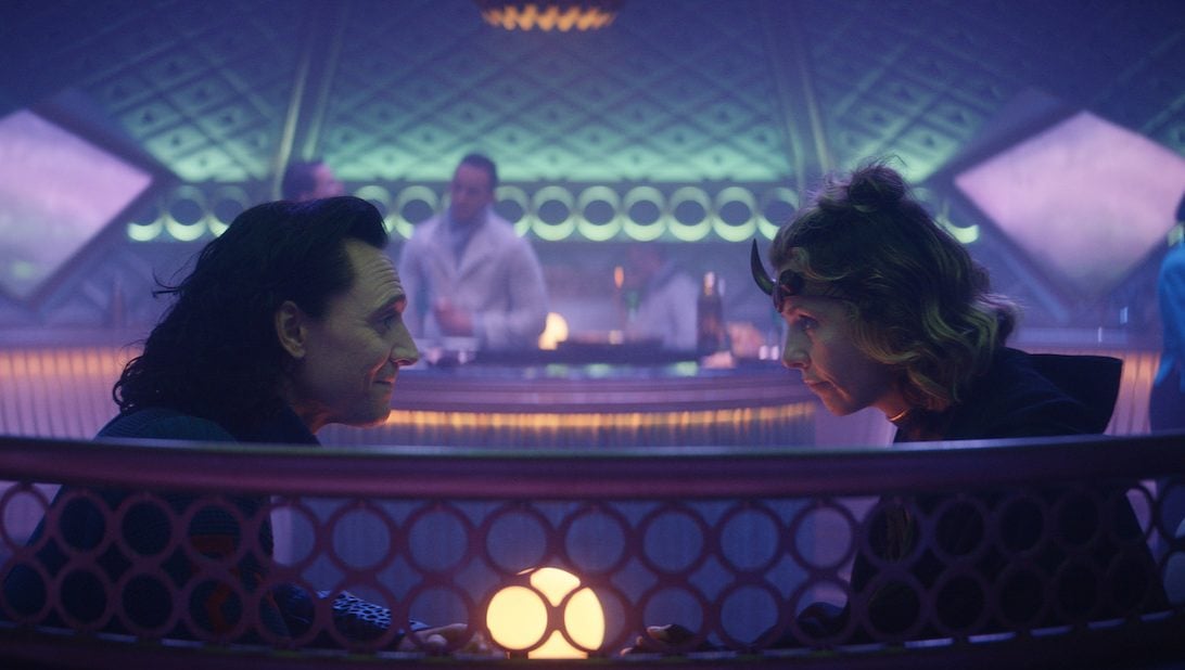 Tom Hiddleston as Loki wearing a blue jumpsuit and sitting across from Sophia Di Martino, who plays Sylvie. She wears a green and gold and black uniform and brass headband with a horn. They sit at a table in a room with purple and blue lights.