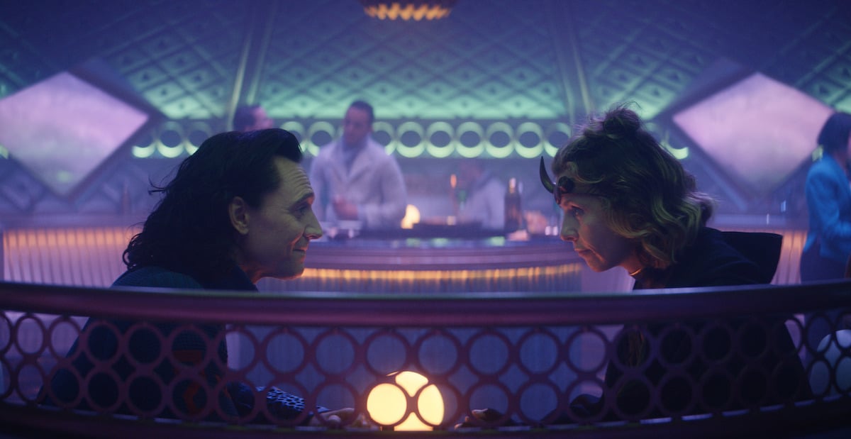 Tom Hiddleston as Loki wearing a blue jumpsuit and sitting across from Sophia Di Martino, who plays Sylvie. She wears a green and gold and black uniform and brass headband with a horn. They sit at a table in a room with purple and blue lights.