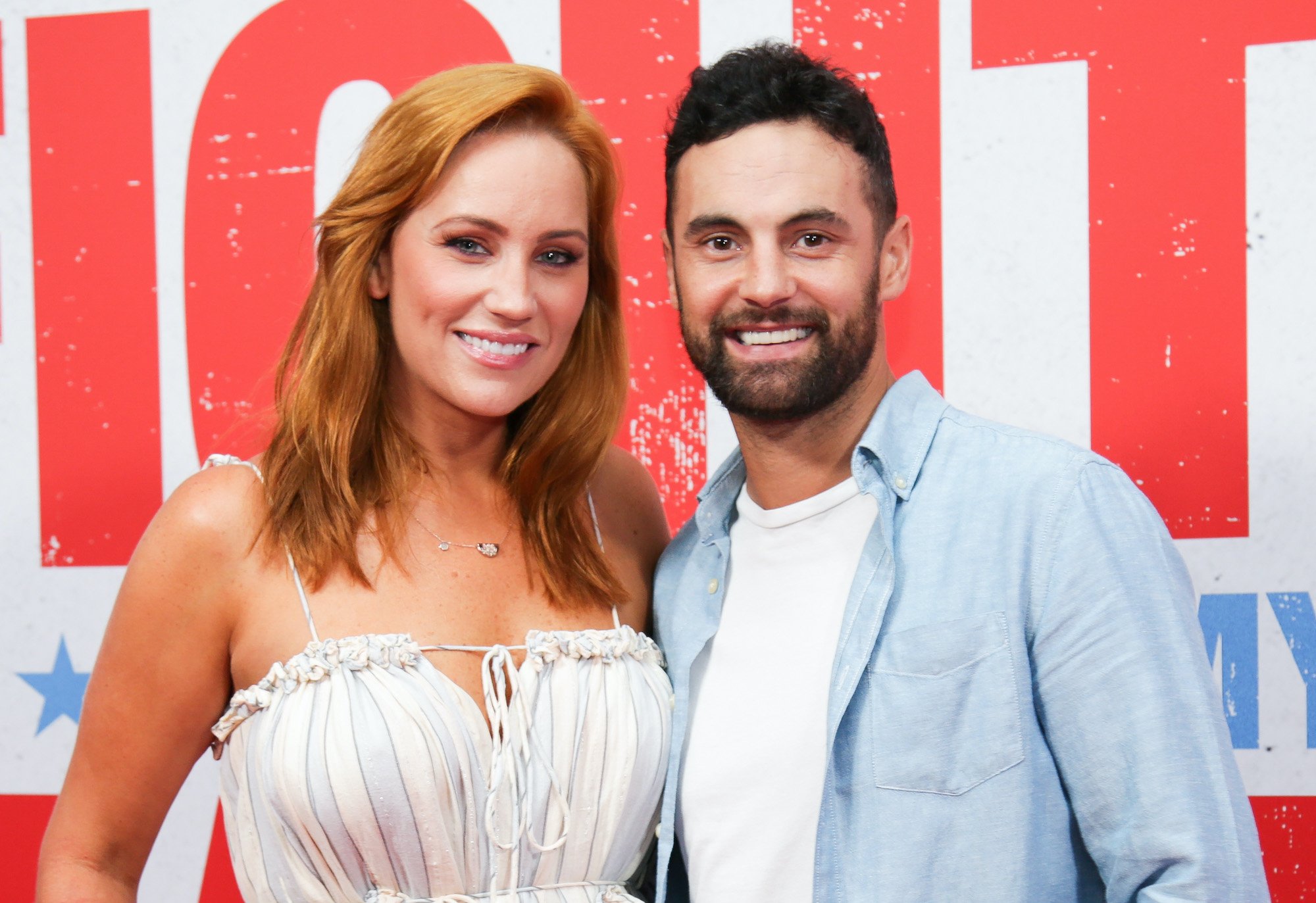 Jules Robinson and Cameron Merchant smiling in front of a red and white background