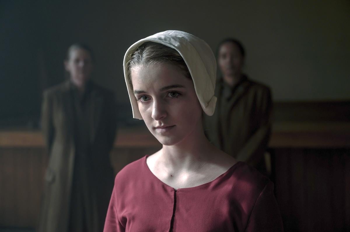 McKenna Grace dressed in a red Handmaid dress and white bonnet as Esther Keyes in 'The Handmaid's Tale' Season 4. Two Aunts dressed in brown dresses and coats stand behind her.