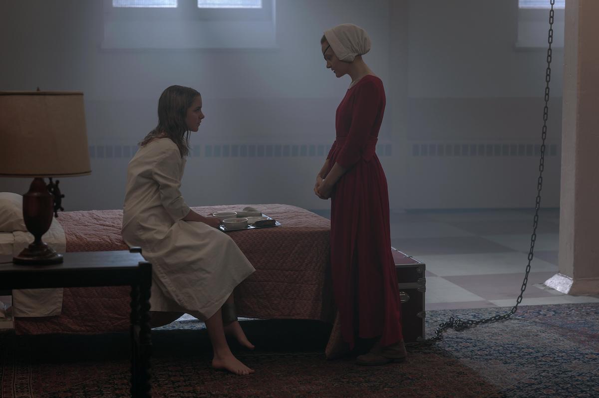 McKenna Grace and Madeline Brewer as Esther and Janine in 'The Handmaid's Tale' Season 4. Grace wears a long-sleeved white nightgown and sits on a bed in a jail cell set up in a former high school gym. Brewer wears a red Handmaid dress and white bonnet.