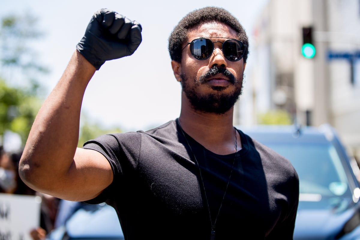 The Wire alum Michael B. Jordan wears black sunglasses, a black T-shirt, and. a black glove as he throws up a fist