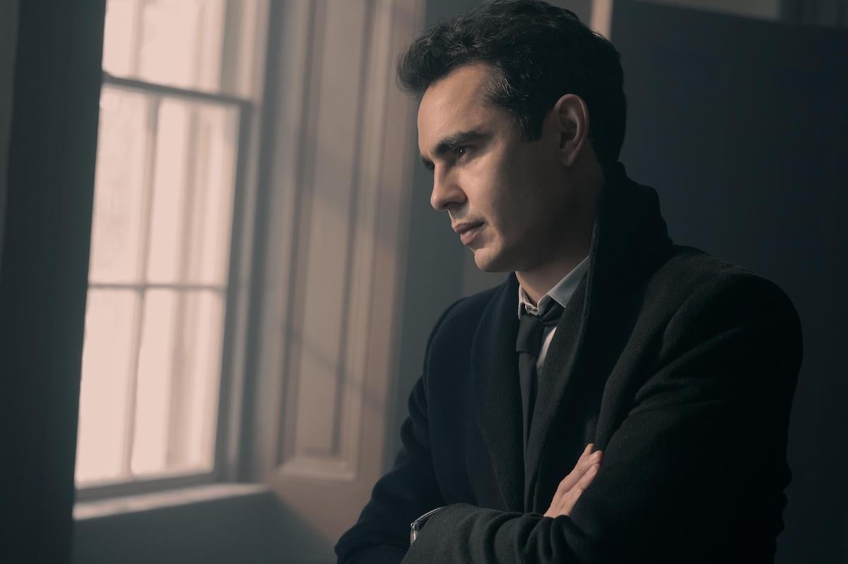 Max Minghella wears a black suit, black wool coat, and crossing his arms as he looks out a window as Nick in 'The Handmaid's Tale' Season 4 Episode 9, 'Progress'