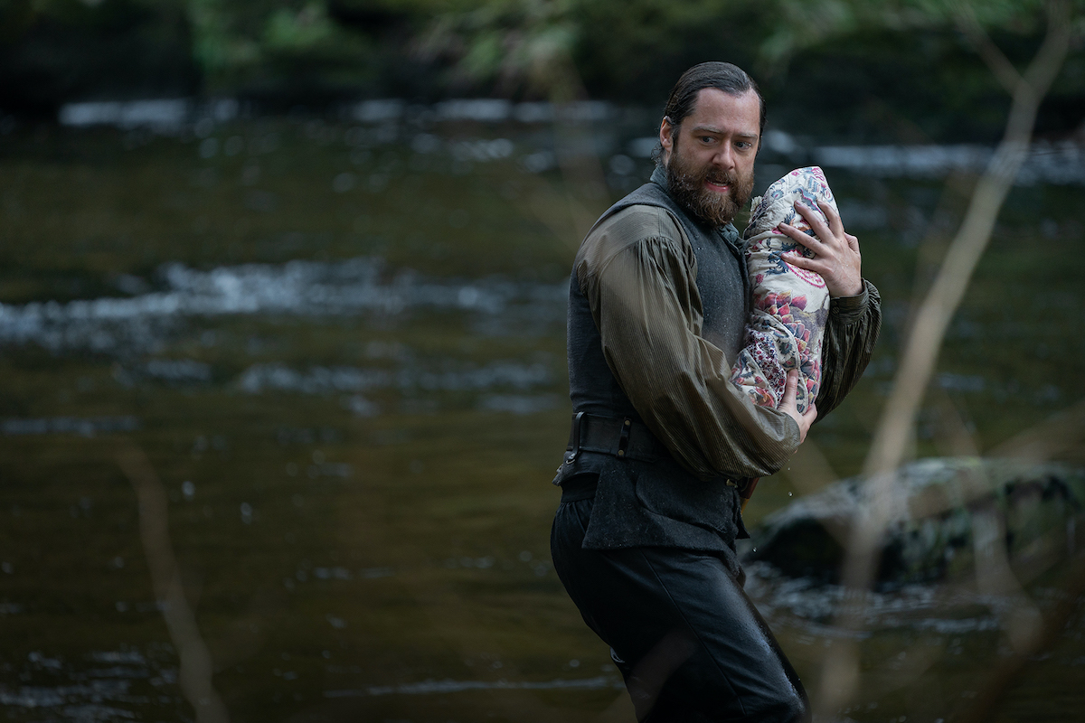 Richard Rankin as Roger in 'Outlander' Season 6 running by a river looking concerned and seemingly carrying a baby wrapped in a quilt