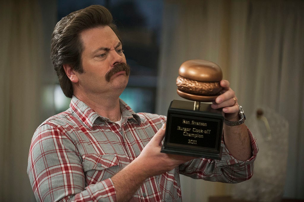 Nick Offerman as Ron Swanson in a plaid shirt holds a golden hamburger statue.