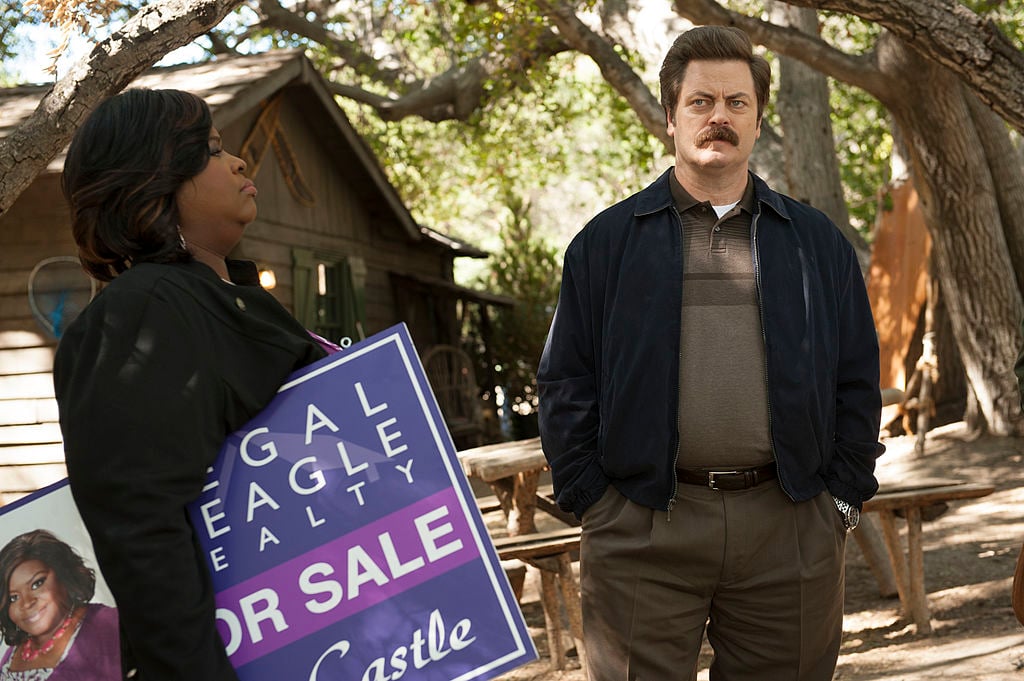 Retta as Donna Meagle holds a real-estate sign while standing next to Nick Offerman as Ron Swanson whose hands are in his pockets.