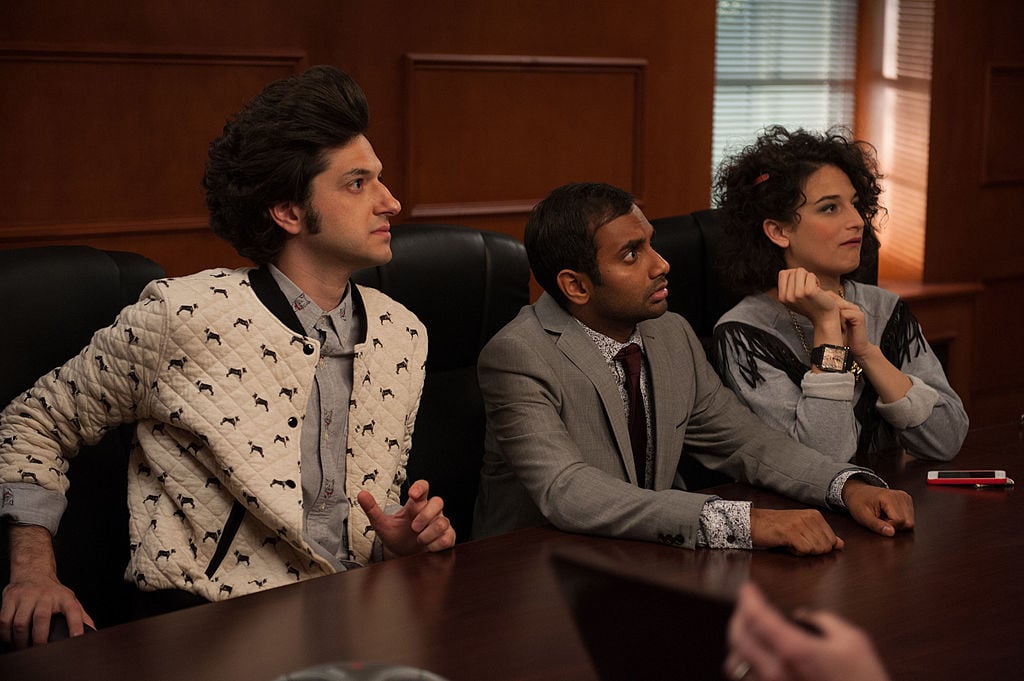 Ben Schwartz as Jean-Ralphio, Aziz Ansari as Tom Haverford, Jenny Slate as Mona Lisa sit at a table together looking at someone speaking who is out of frame.