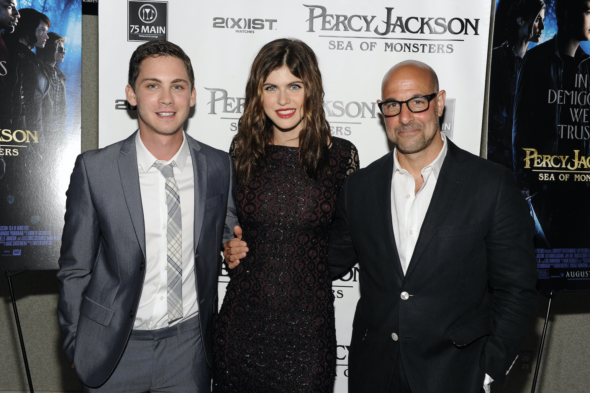 Logan Lerman, Alexandra Daddario and Stanley Tucci smiling in front of a 'Percy Jackson' banner