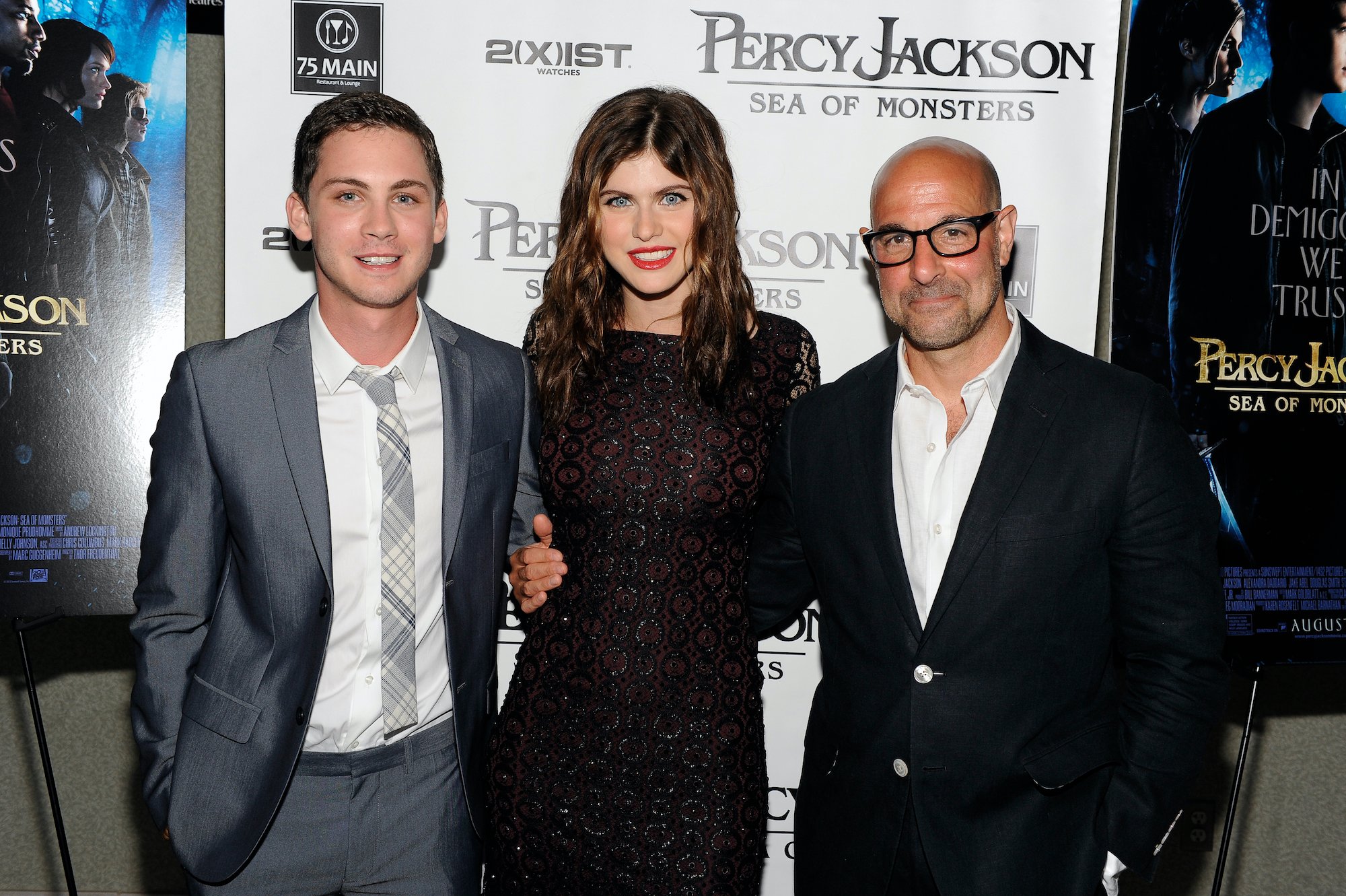 Logan Lerman, Alexandra Daddario and Stanley Tucci smiling in front of a 'Percy Jackson' banner