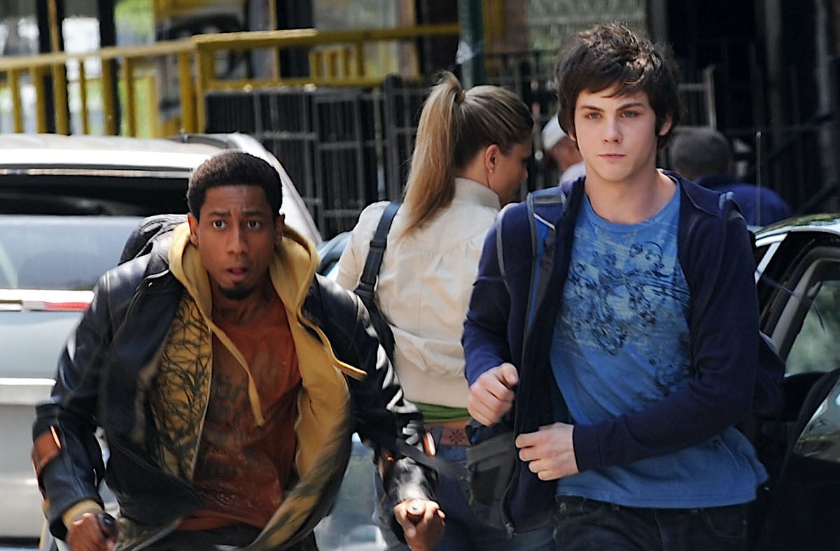 Logan Lerman dressed in a blue shirt and blue hoodie and jeans as Percy Jackson. He runs through a street in Brooklyn, New York with cast mate Brandon T. Jackson, who played Grover. Jackson wears an orange T-shirt, yellow zip-up hoodie, and black leather jacket.