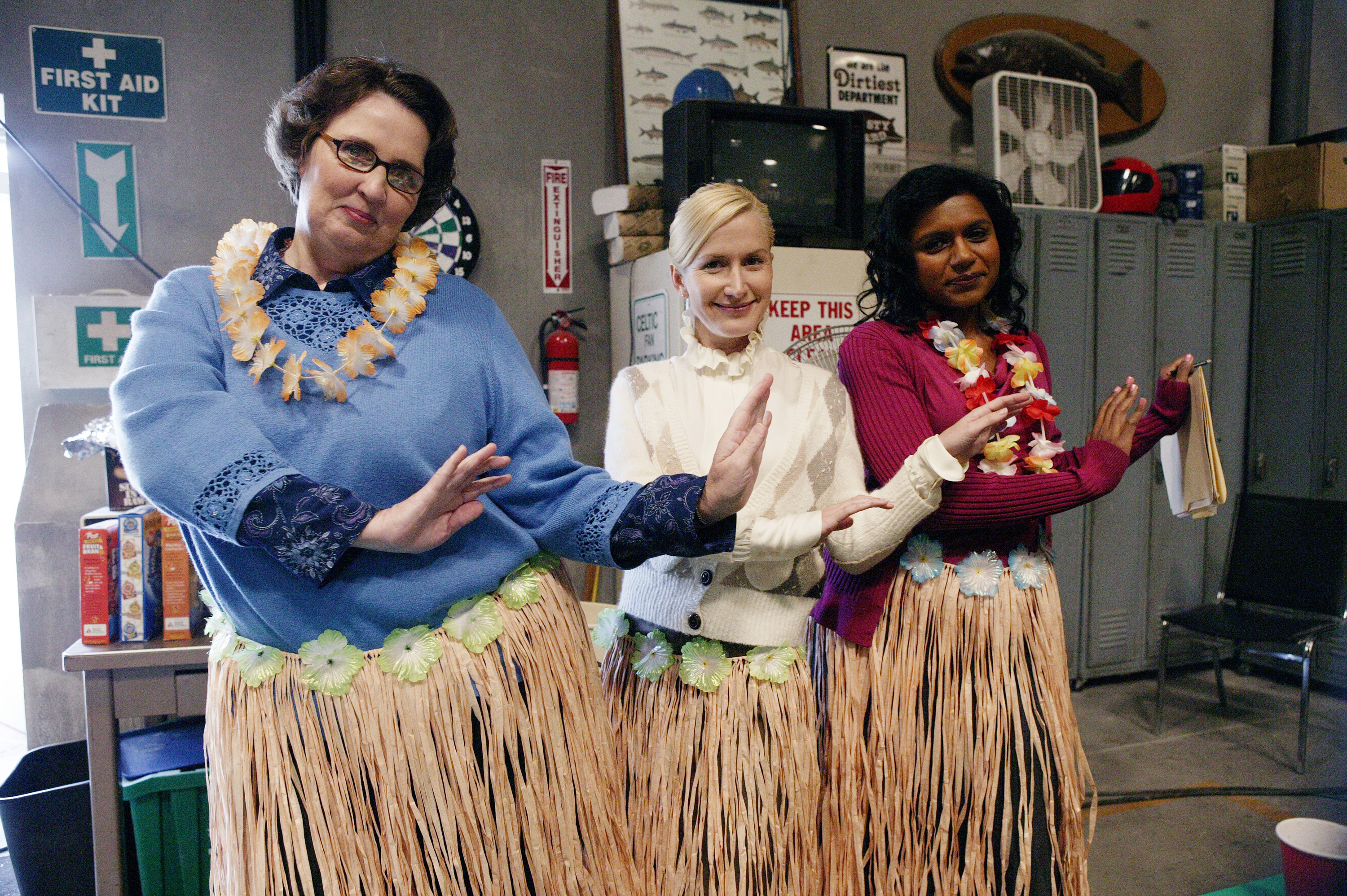 The Office cast members Phyllis Lapin, Angela Kinsey, and Mindy Kaling as Phyllis Smith, Angela Martin, and Kelly Kapoor wearing hula skirts while filming an episode