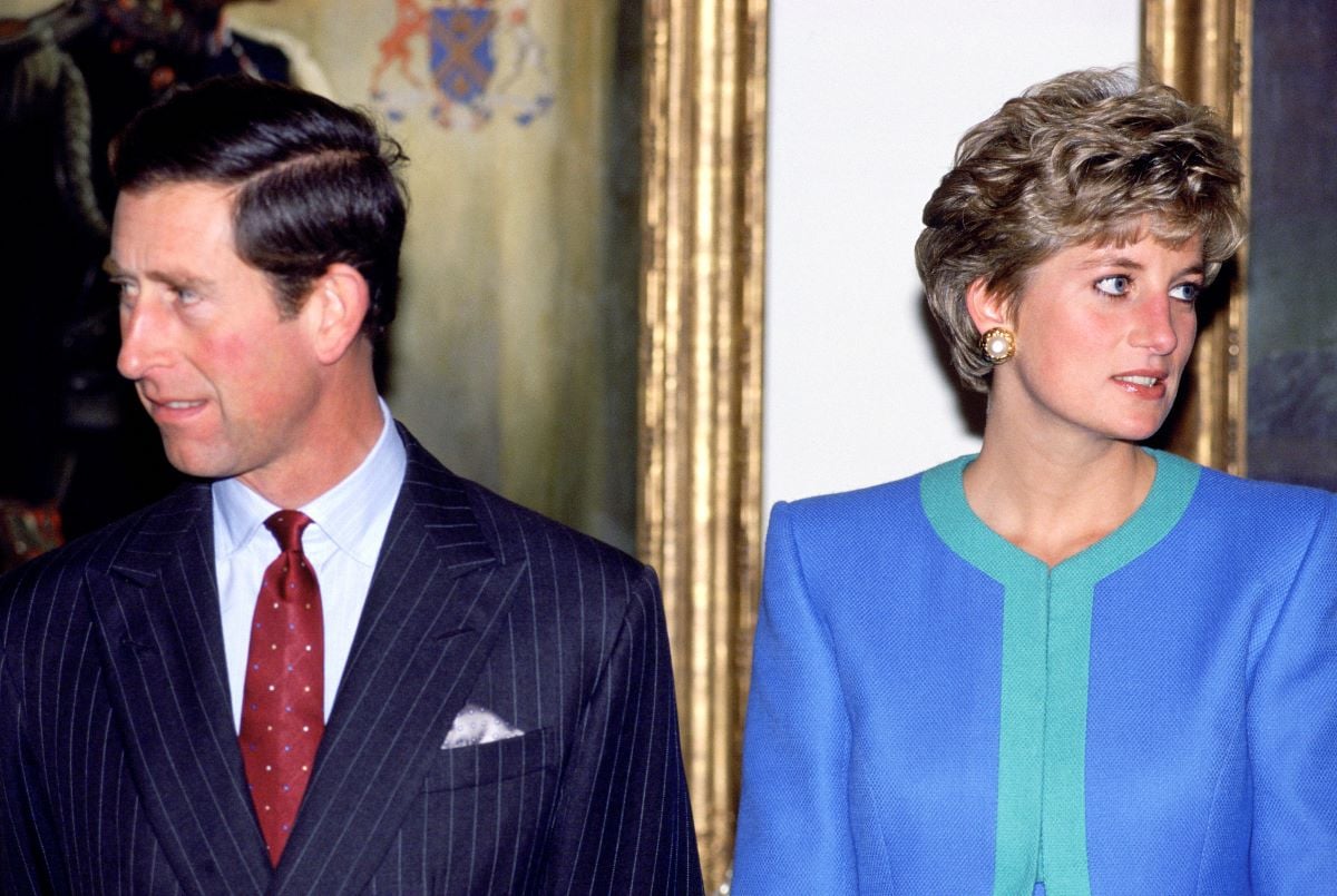 Prince Charles in a navy suit with a burgundy tie looking away from Princess Diana who is in a blue suit
