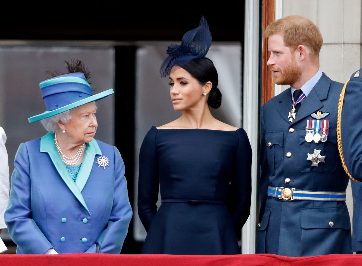 Queen Elizabeth II in a blue hat and suit, Meghan Markle in a navy dress and hat, and Prince Harry in military attire