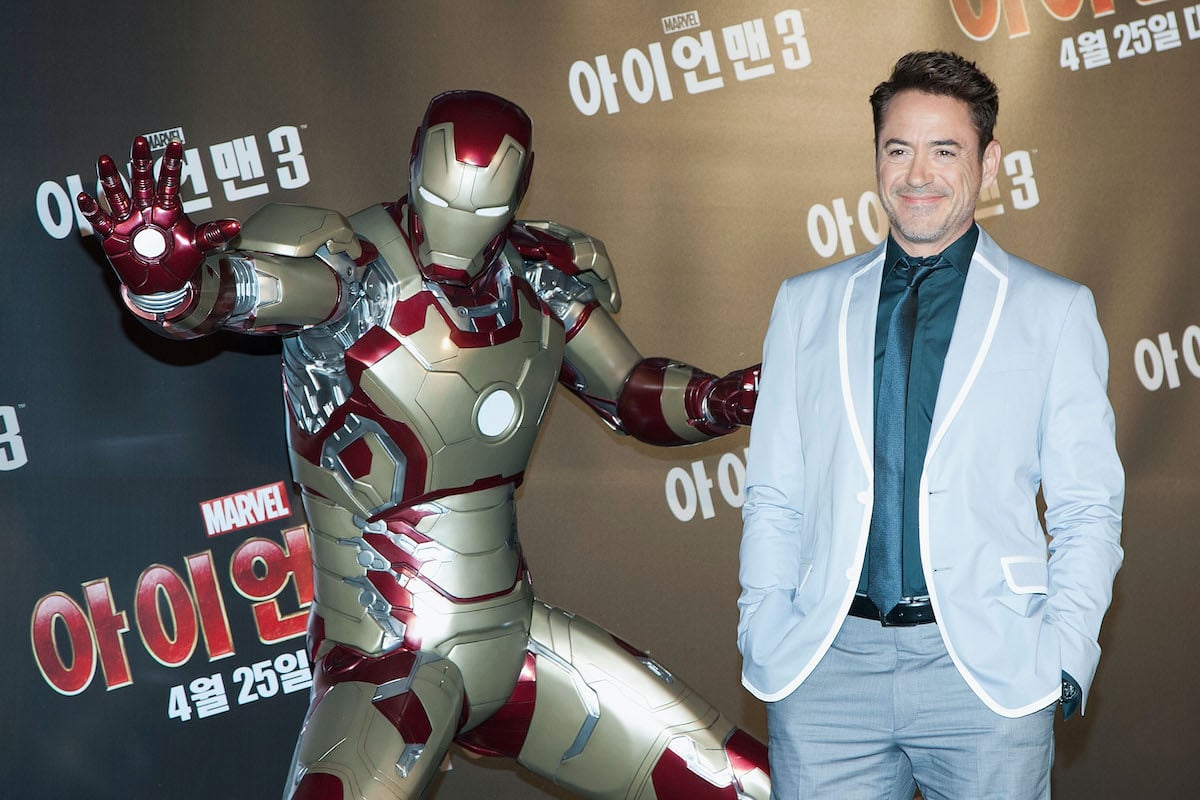 Robert Downey Jr. wears a suit and smiles while posing next to an Iron Man suit