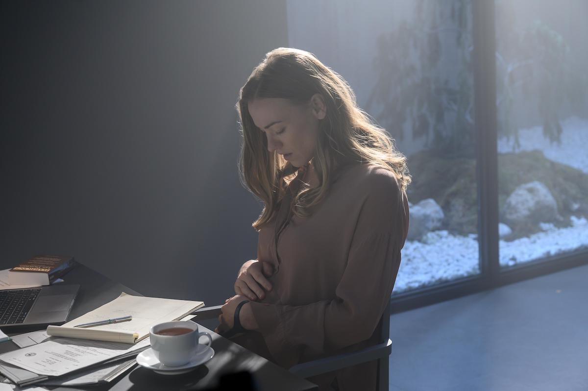 Yvonne Strahovski as Serena Joy Waterford in 'The Handmaid's Tale' Season 4. She sits at a desk covered in papers, a laptop, and a full cup of tea in her prison cell. Serena wears a light pink blouse and looks down as she cradles her baby bump, a concerned look on her face.