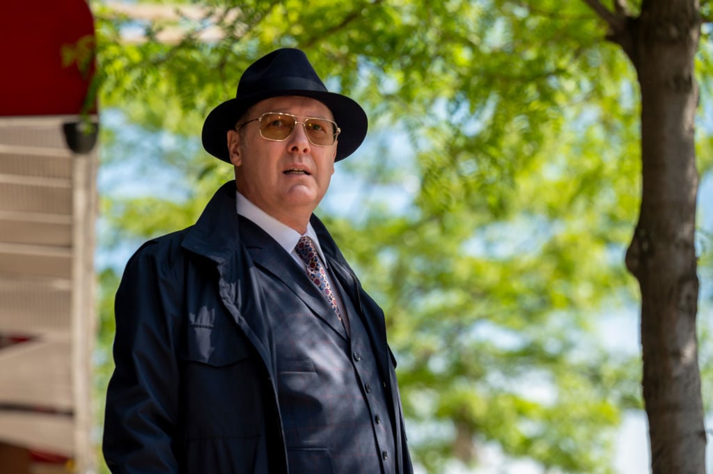 James Spader as Raymond "Red" Reddington looks into theopen air. He's dressed in a navy trench coat, his trademark hat, and sunglasses.