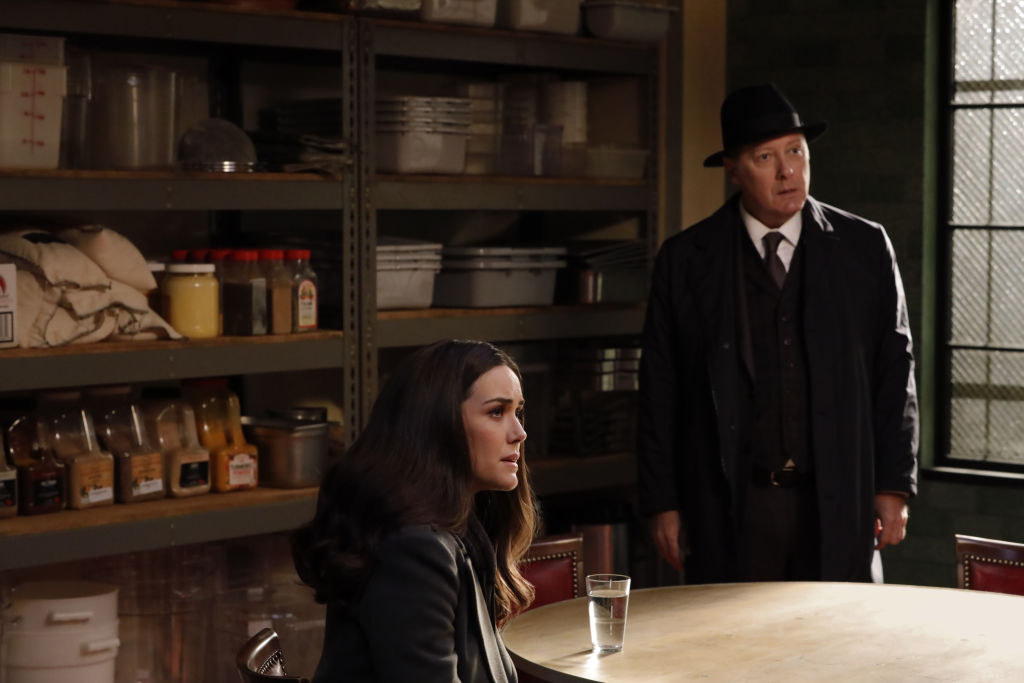 Megan Boone as Liz Keen sits at a table while James Spader as Raymond Reddington stands next to her.