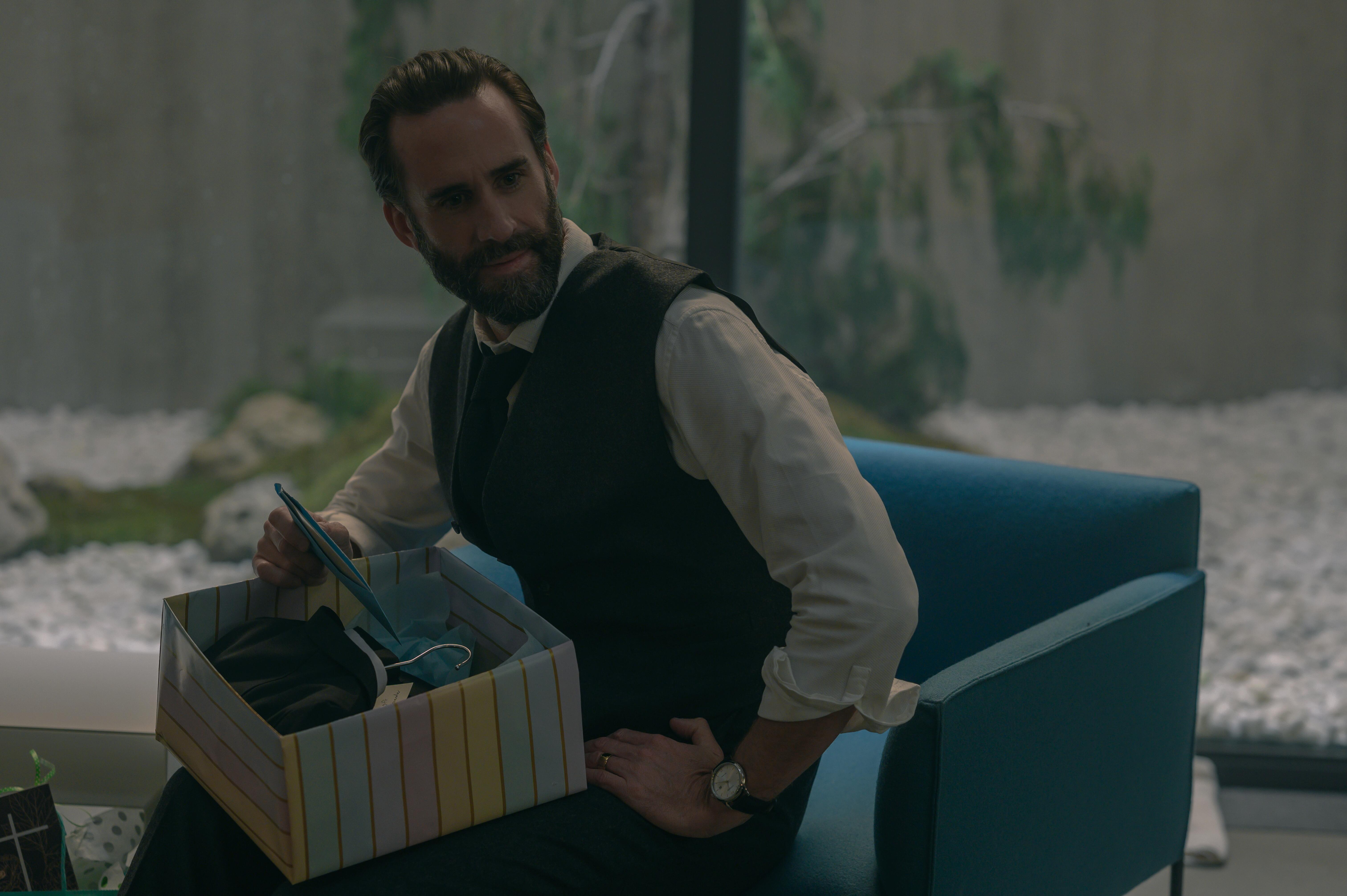 Fred Waterford sits in a blue chair with a wrapped present in his lap in 'The Handmaid's Tale'