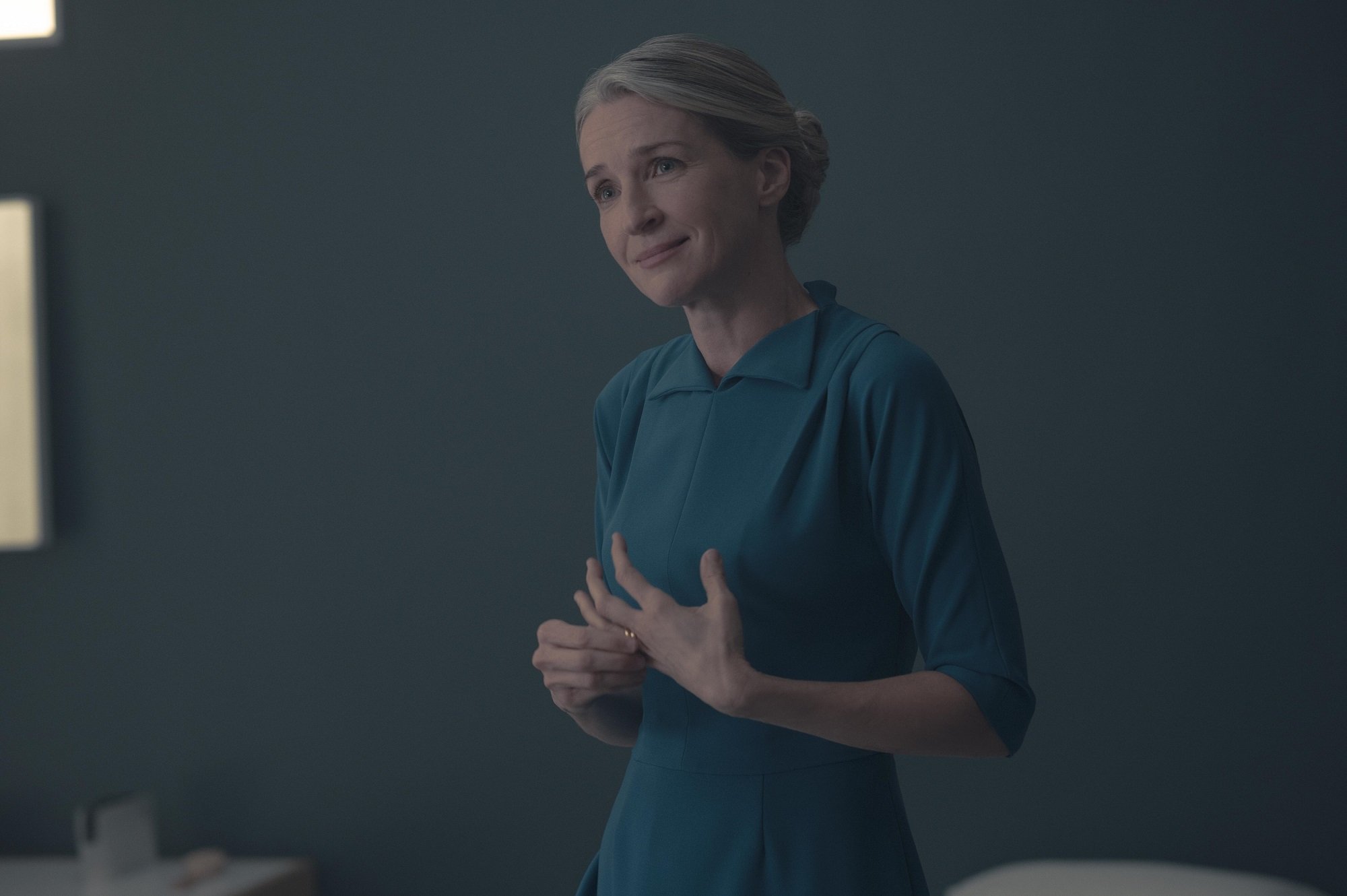 Naomi Putnam in her blue Wives' dress stands in front of a gray wall in 'The Handmaid's Tale'