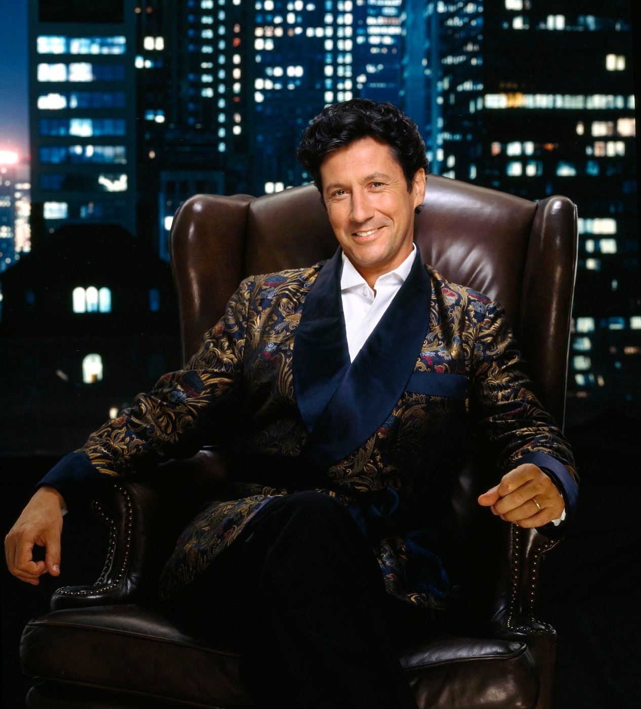 Charles Shaughnessy as Maxwell Sheffield is photographed sitting in a leather chair against a backdrop of the New YorK City skyline