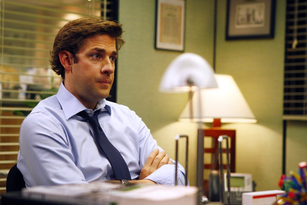 John Krasinski as Jim Halpert sits with his arms crossed at Michael Scott's desk.