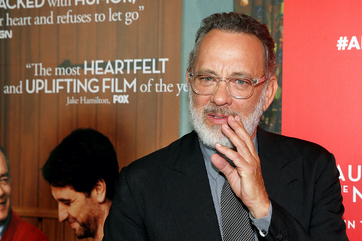 Tom Hanks wears a suit and tie and stands in front of a display for 'A Beautiful Day In The Neighborhood'