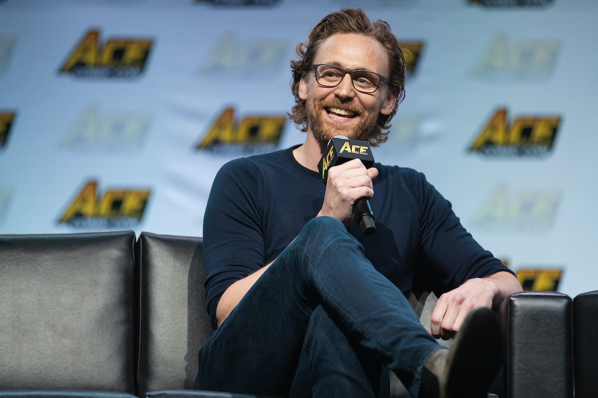 Tom Hiddleston sits on a dark sofa holding a microphone and smiling