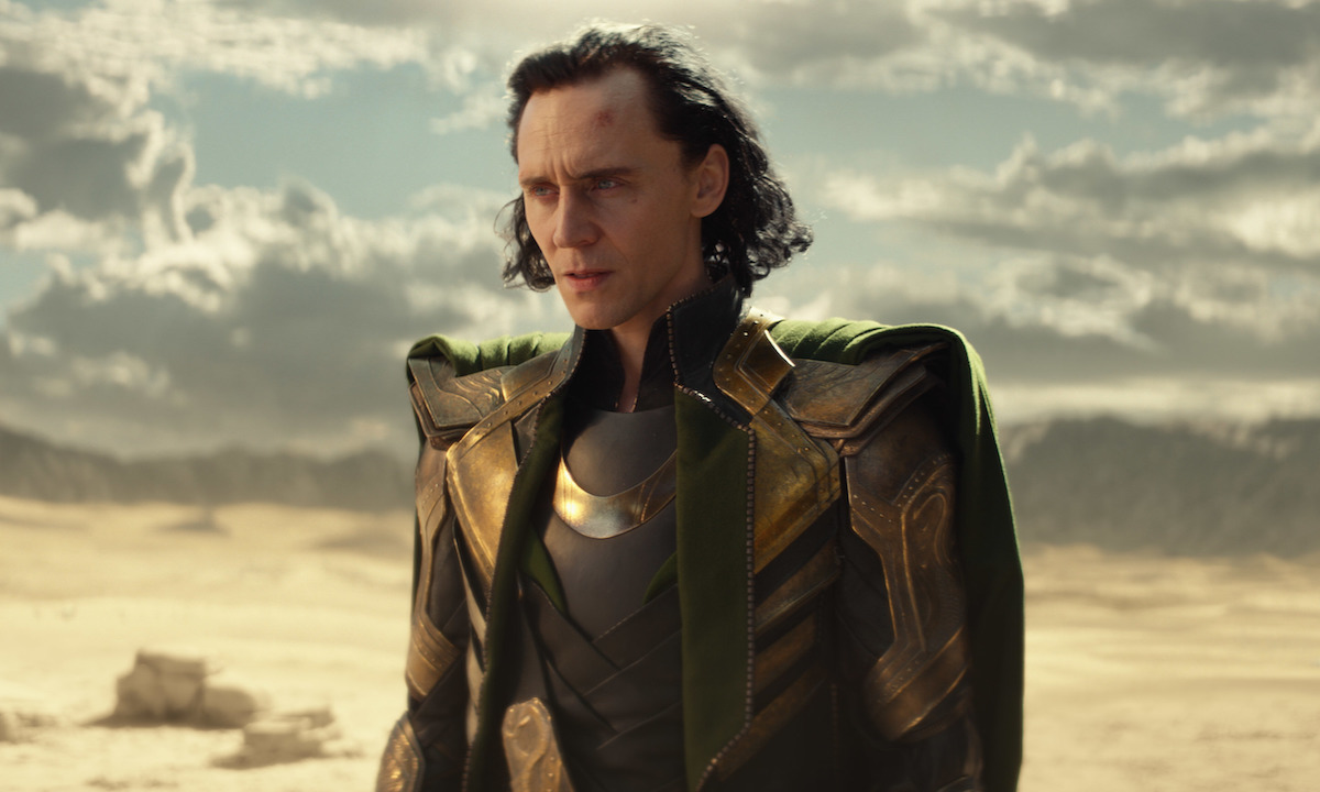 Tom Hiddleston in grey and gold armor with a green cape while standing in a desert as Loki in 'Loki' on Disney+