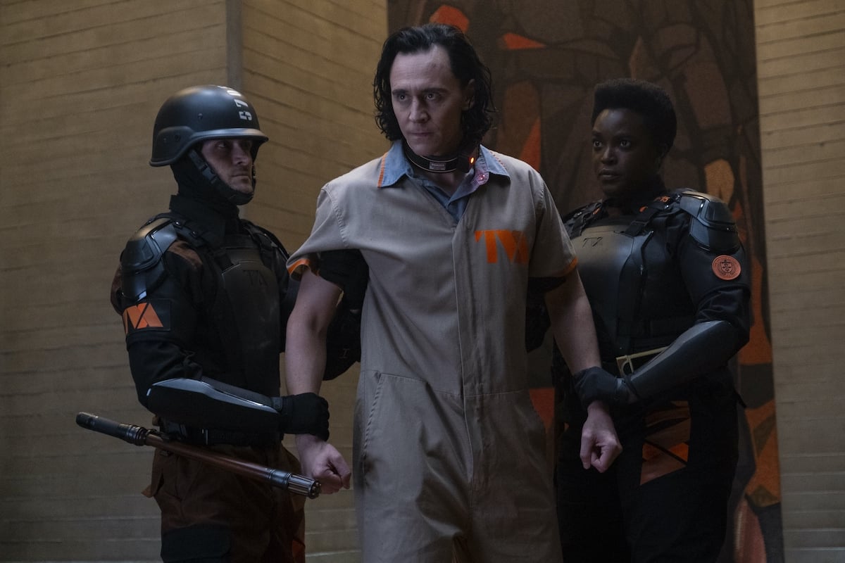 Tom Hiddleston as Loki in a beige and orange prison uniform is held back by Wunmi Mosaku as Hunter B-15 and another officer in all-black uniforms in 'Loki' on Disney+.